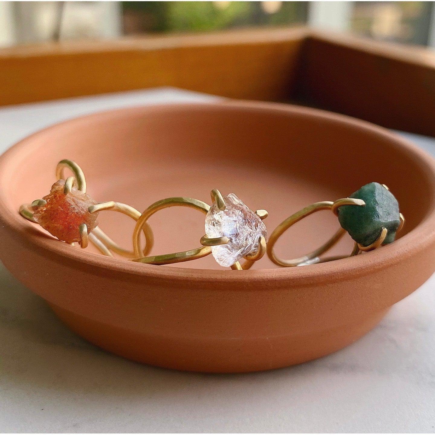 raw gemstone rings with brass double wire band with sunstone which is an orange gemstone, ametrine which is clear to purple, and emerald which is a forest green color. The rings are sitting in a terracotta pottery dish.