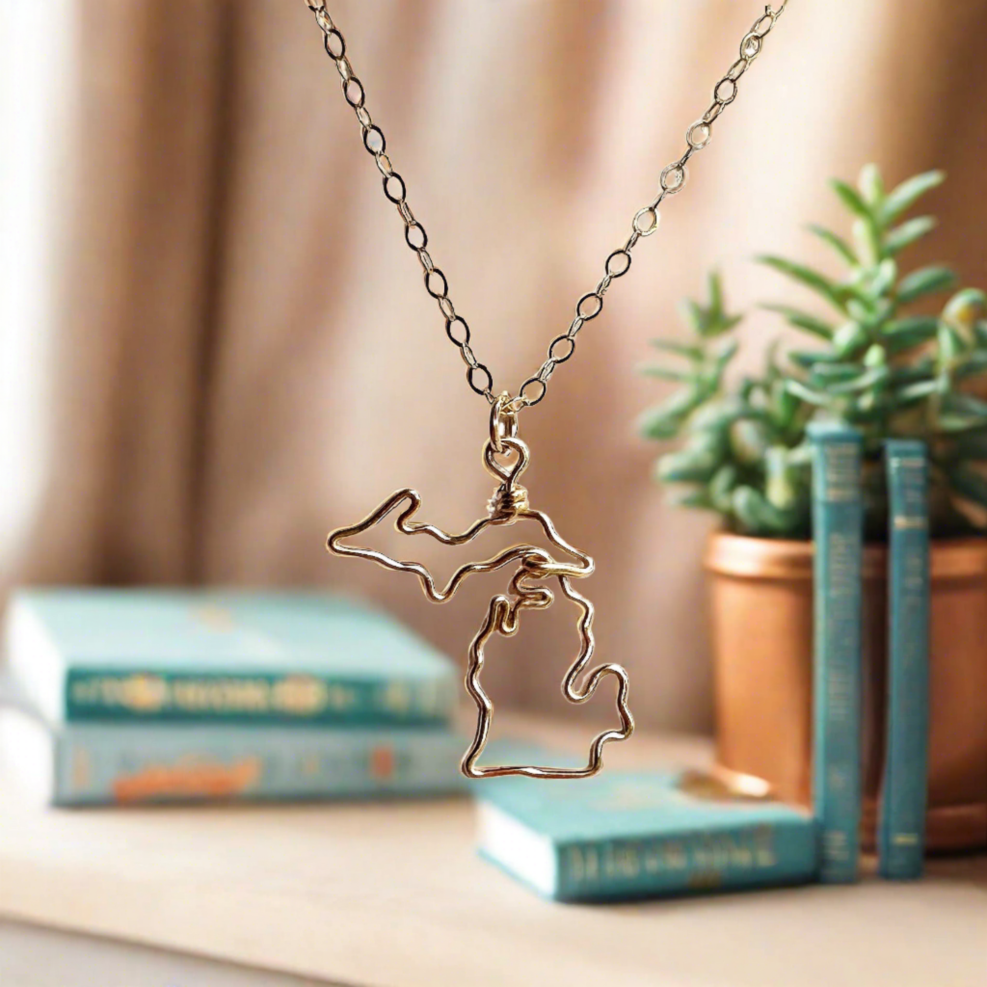 gold Michigan necklace with books and plants and sunlight in the background 